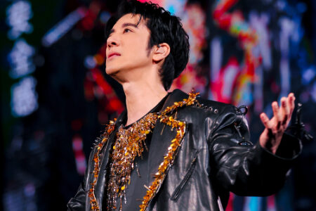 Leehom performing on stage in an ornate black leather jacket with gold embellishments, looking upward with colorful stage lights