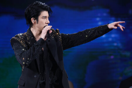Leehom in a black sequined blazer pointing dramatically while performing with blue and green stage lighting
