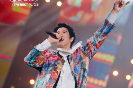 Leehom performing energetically in a vibrant colorful tie-dye jacket with one arm raised on stage