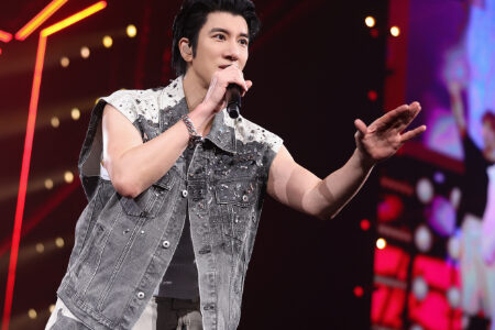 Close-up of Leehom in denim vest holding microphone with warm bokeh stage lights behind him