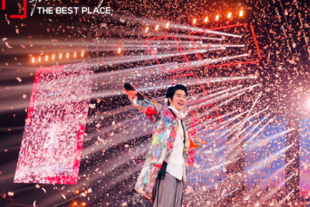 Leehom in colorful tie-dye jacket with arm raised triumphantly surrounded by dramatic water spray effects on stage